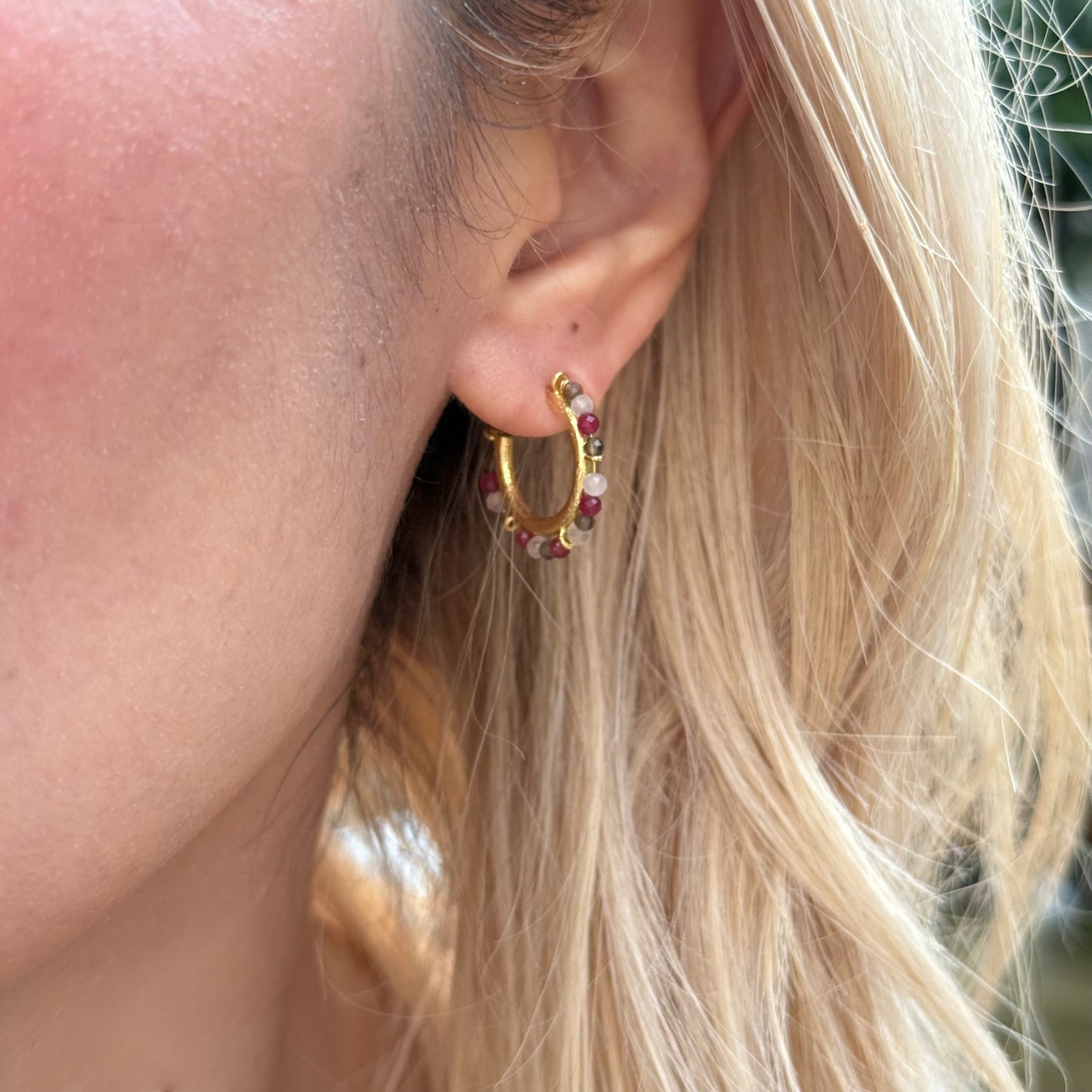 Close-up of an ear wearing a gold hoop earring with red gemstones, blonde hair.