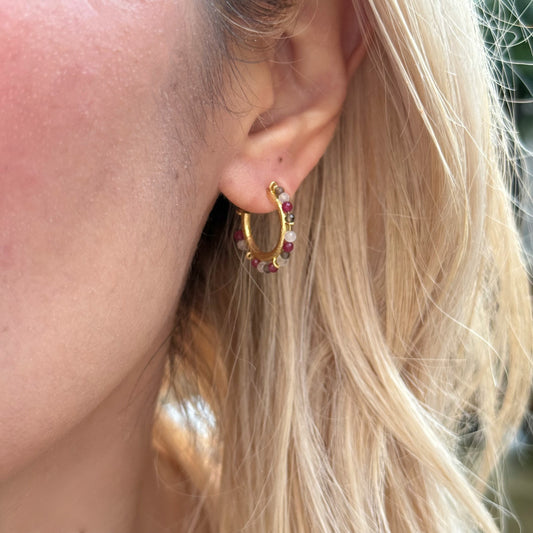 Close-up of an ear wearing a gold hoop earring with red gemstones, blonde hair.
