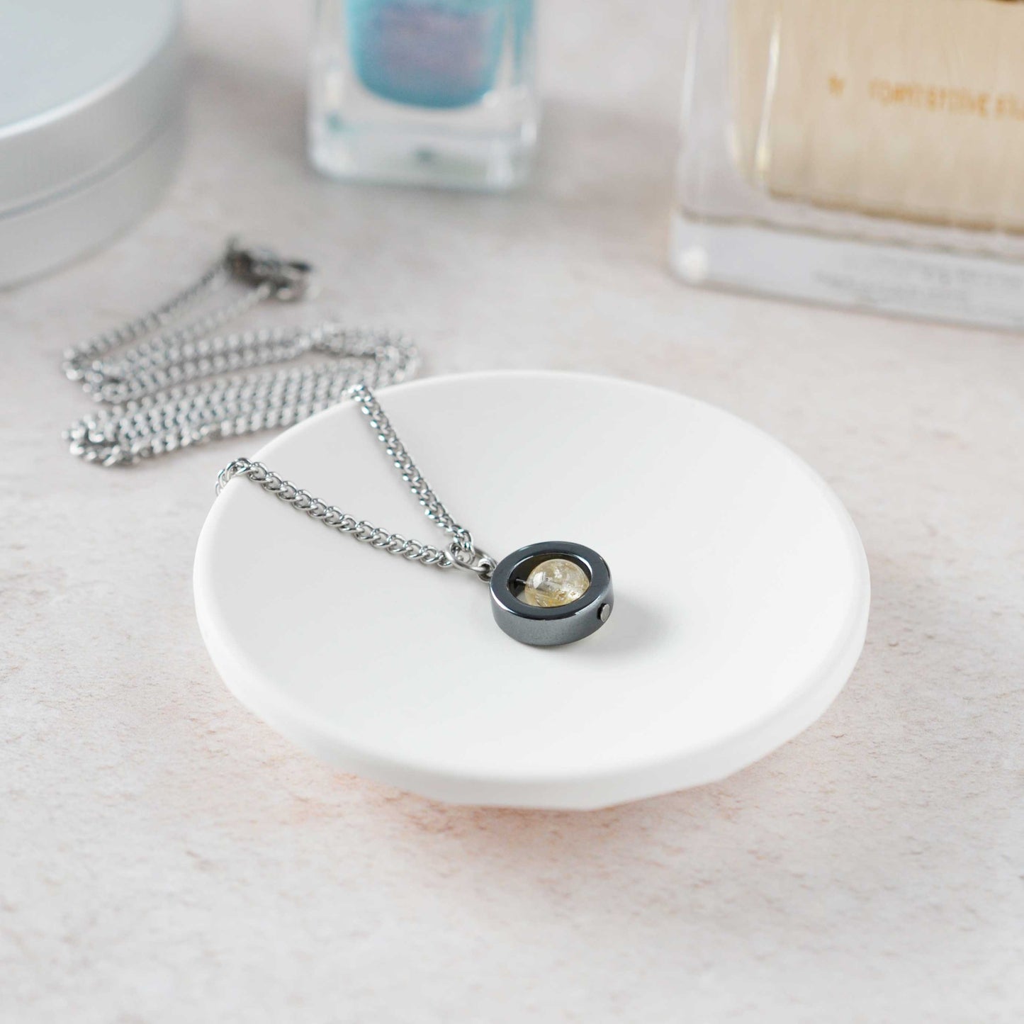 Necklace with a pendant on a white dish, with nail polish and a bottle in the background.