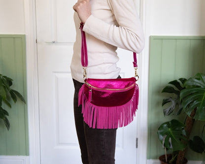 A person wearing a white top is holding a raspberry metallic leather half moon fringed bag with a pink fringe and gold hardware.