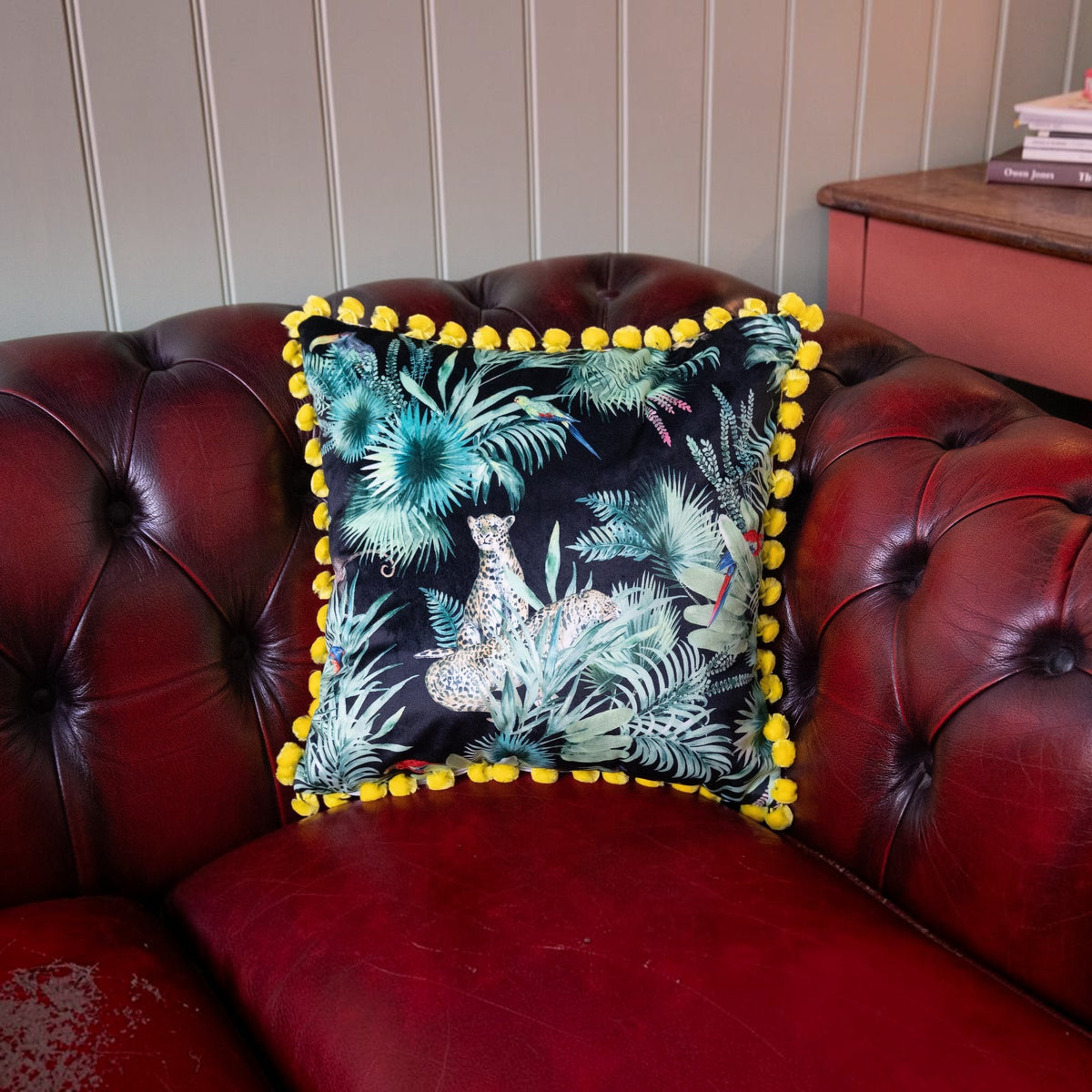 Deepest Jungle velvet cushion with acid yellow pompoms on a deep red leather Chesterfield sofa