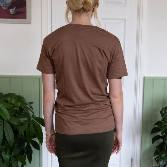 Person wearing a brown t-shirt standing indoors with plants in the background