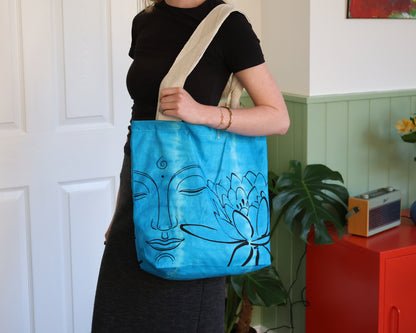A person wearing a black t-shirt and dark skirt carries a bright blue canvas tote bag with a beige shoulder strap. The bag features a black line art design of a face and a lotus flower.