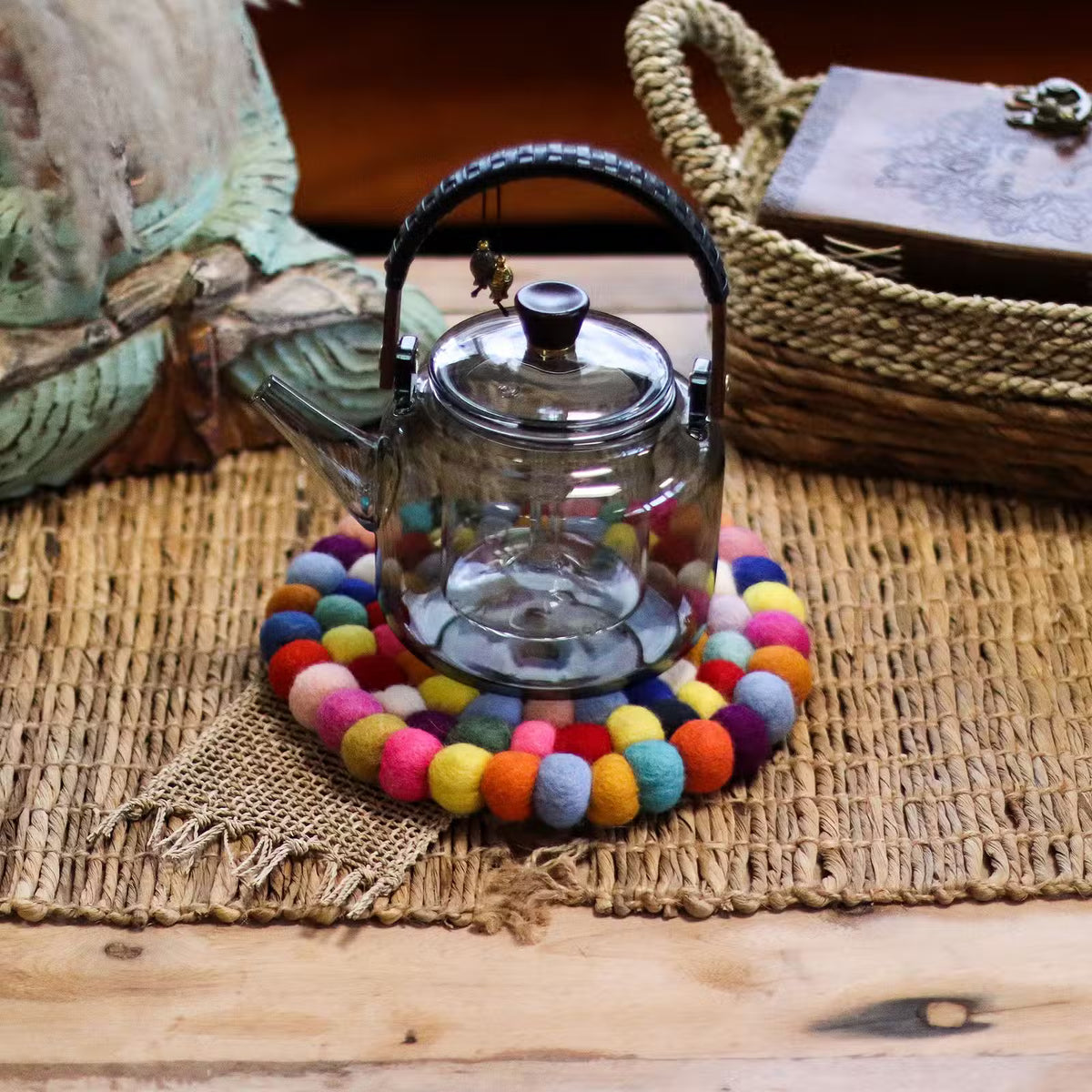 Multicolour felt ball round coaster under a glass teapot on a woven natural mat