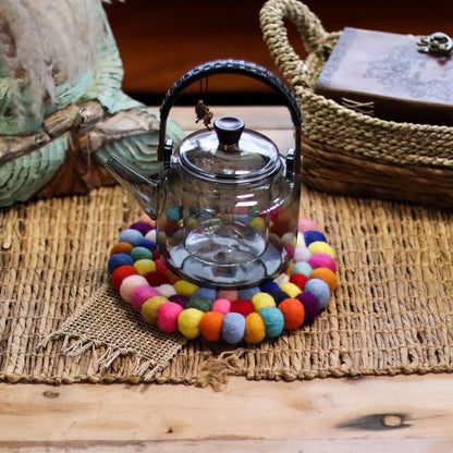 Multicolour felt ball round coaster under a glass teapot on a woven natural mat