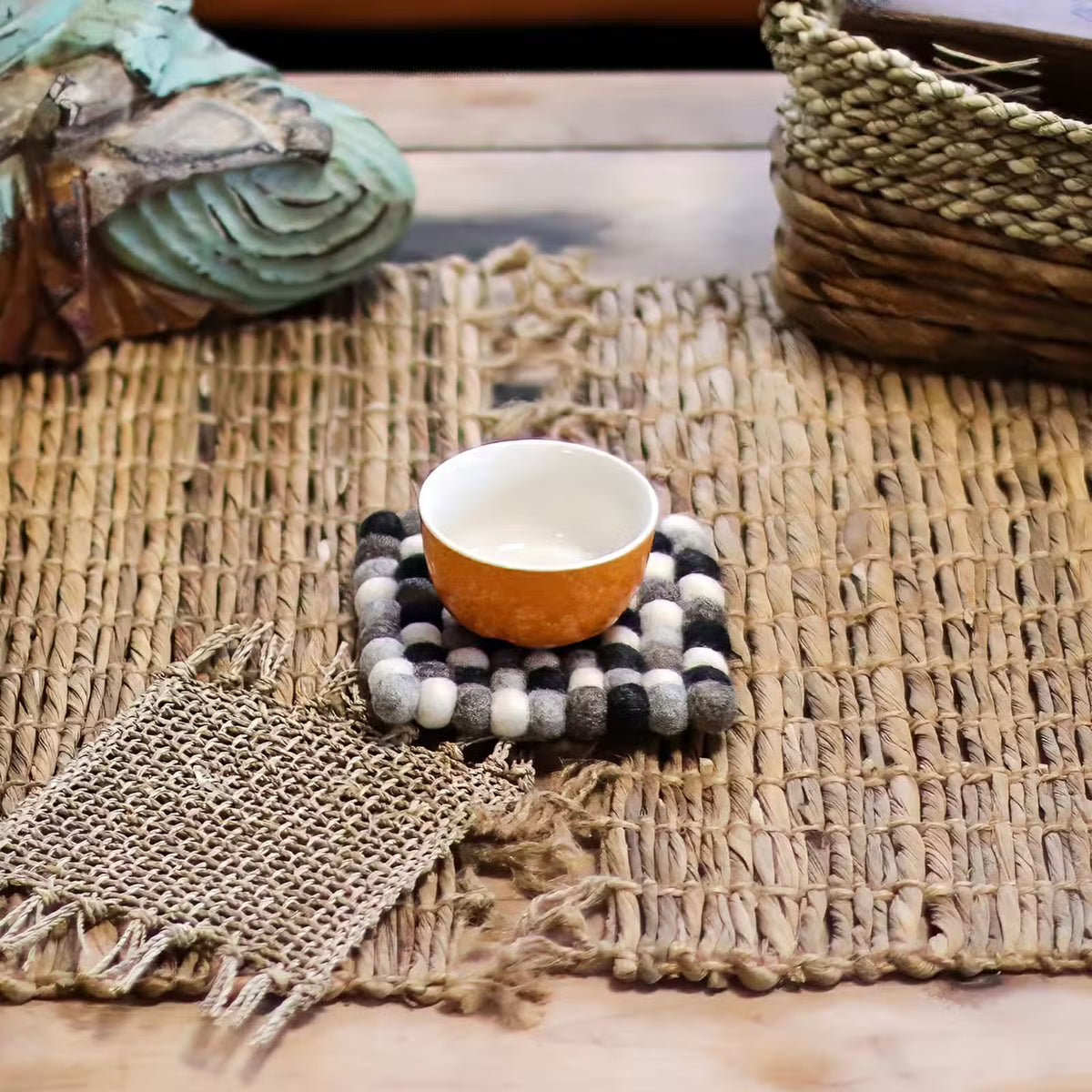 Grey felt ball square coaster under an orange cup on a woven natural mat