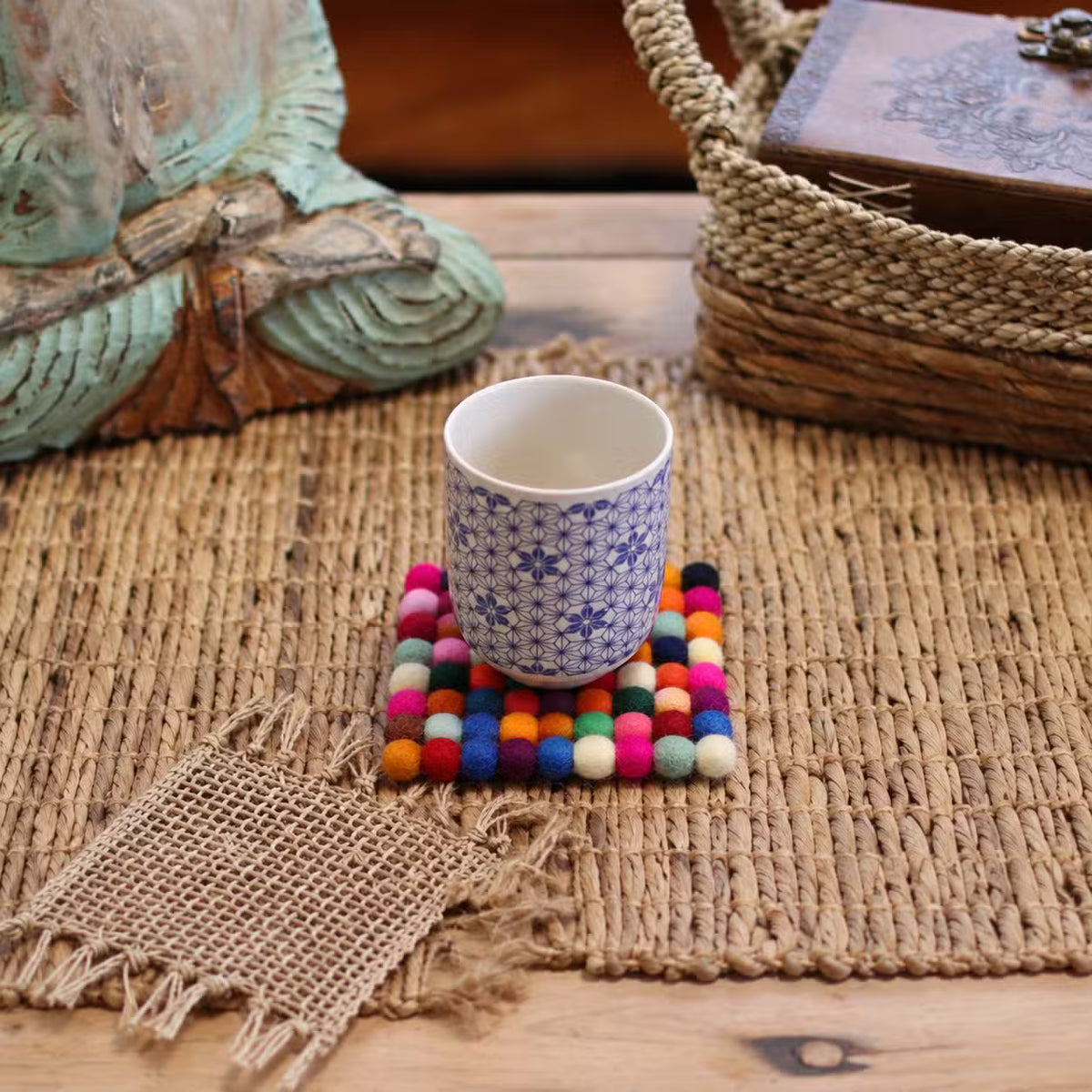 Multicolour felt ball square coaster under a blue and white patterned cup on a woven natural mat
