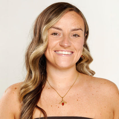 Woman with long wavy hair wearing a gold necklace with a red pendant on a plain background
