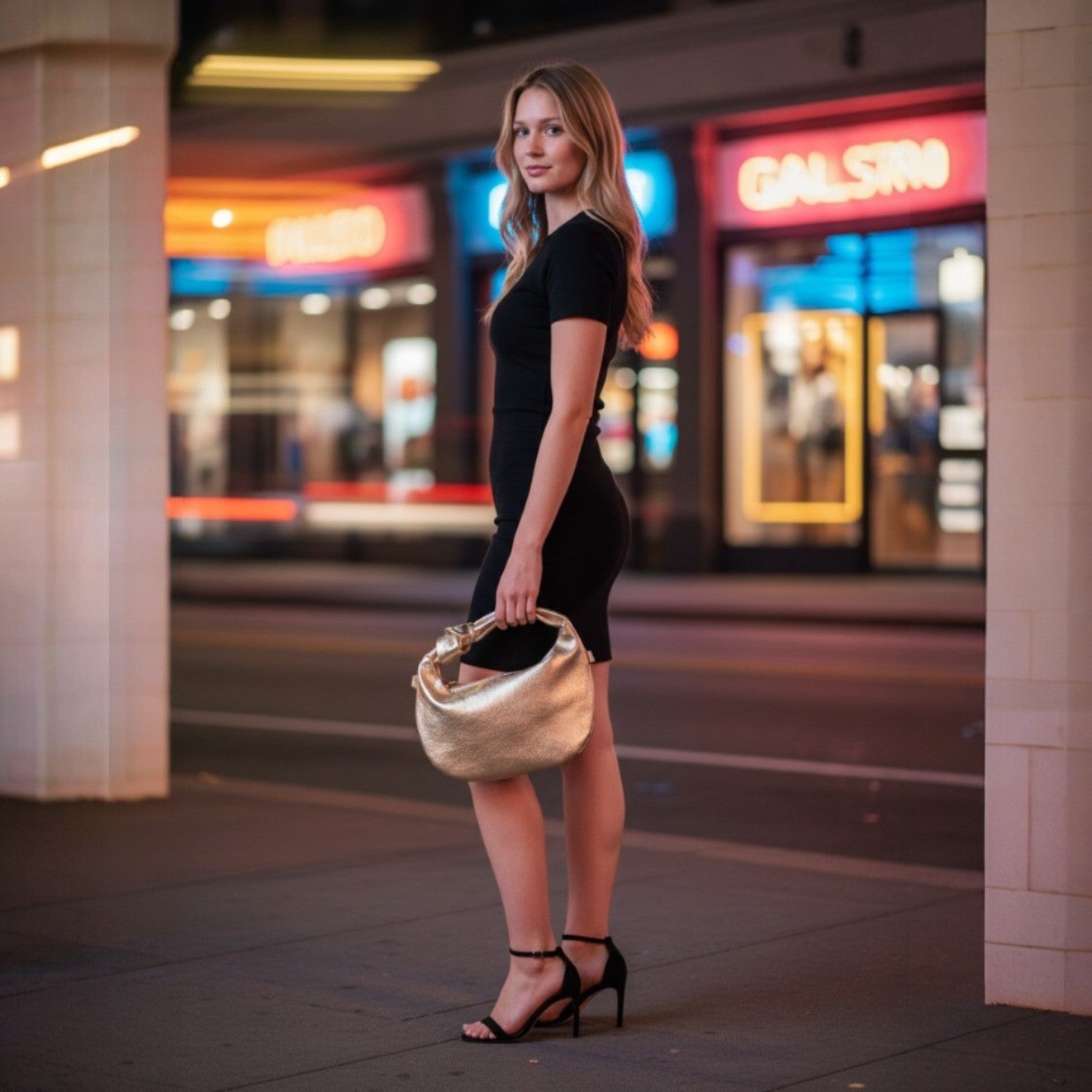 Woman in a black dress holding a gold handbag on a city street at night.