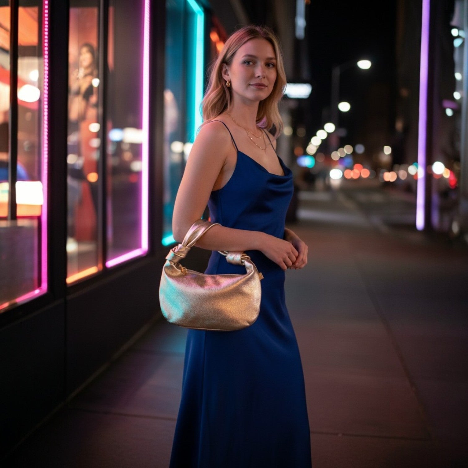 Woman in a blue dress holding a gold handbag in an urban night setting with colorful lights.