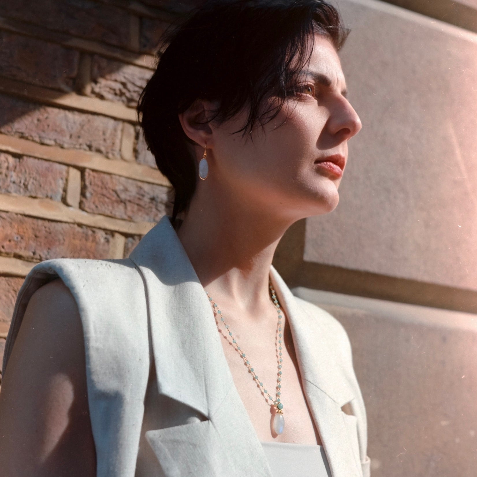 Woman wearing a light gray blazer with a necklace, standing against a stone wall.
