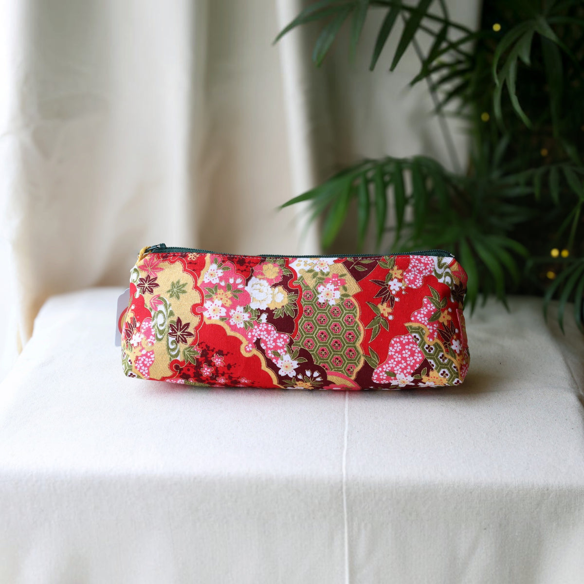 Isumi Red Japanese pencil case with a dark green zip on a cream cloth beside a palm plant