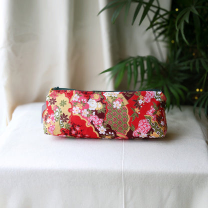 Isumi Red Japanese pencil case with a dark green zip on a cream cloth beside a palm plant