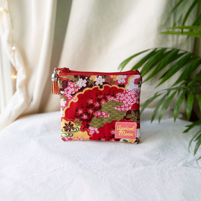 Isumi Red Japanese purse with a red zip and orange tassel on a cream cloth beside a palm plant