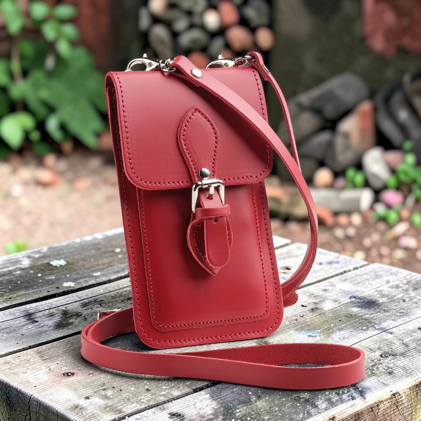 Red leather crossbody bag on a wooden surface with a garden background