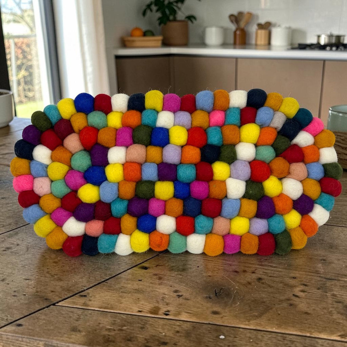 Multicolour felt ball oval centre mat standing on a rustic wooden surface in a kitchen setting