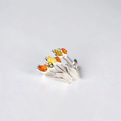 A silver brooch designed as a cluster of tulips, featuring faceted amber gemstones in amber orange, bright yellow, and olive green, set against a plain light grey background.
