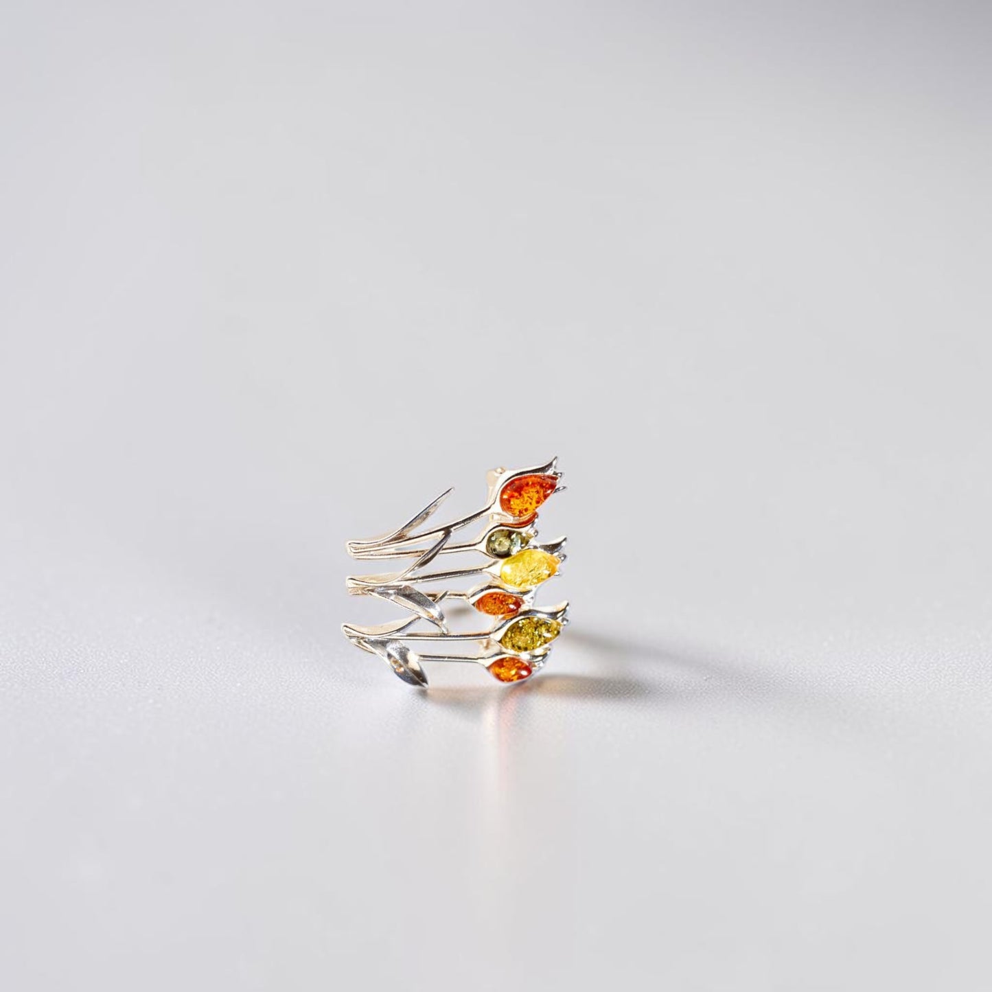 A delicate silver brooch featuring multiple thin branches adorned with small marquise-cut amber gemstones in orange, yellow, and green, photographed on a clean light grey surface.
