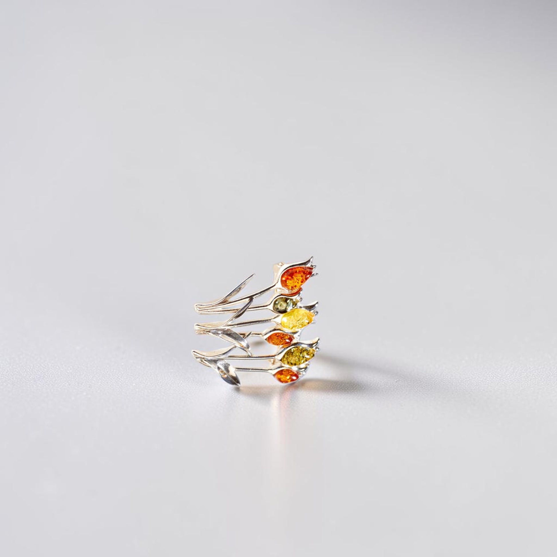 A delicate silver brooch featuring multiple thin branches adorned with small marquise-cut amber gemstones in orange, yellow, and green, photographed on a clean light grey surface.