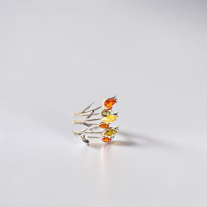 A delicate silver brooch featuring multiple thin branches adorned with small marquise-cut amber gemstones in orange, yellow, and green, photographed on a clean light grey surface.