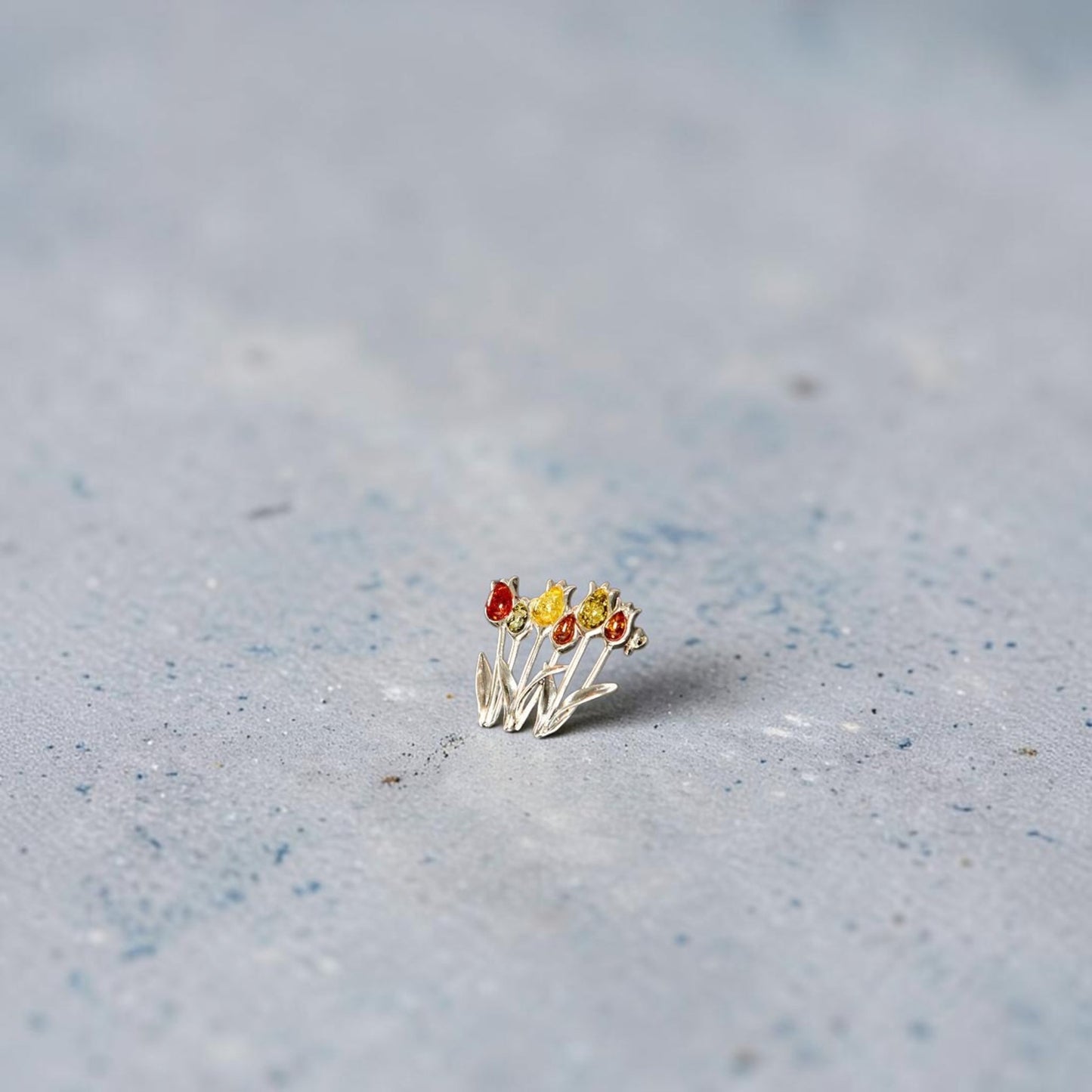 A delicate sterling silver brooch featuring three small tulip flowers with red, yellow, and orange amber petals, resting on a textured light grey surface.