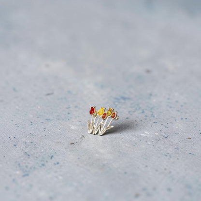 A delicate sterling silver brooch featuring three small tulip flowers with red, yellow, and orange amber petals, resting on a textured light grey surface.