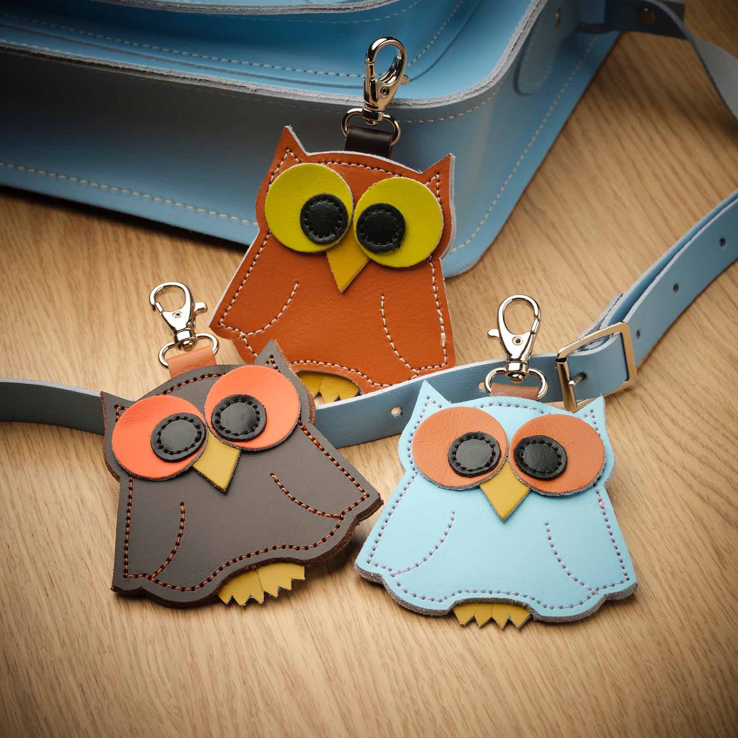 Three owl-shaped keychains in brown, gray, and blue on a wooden surface.