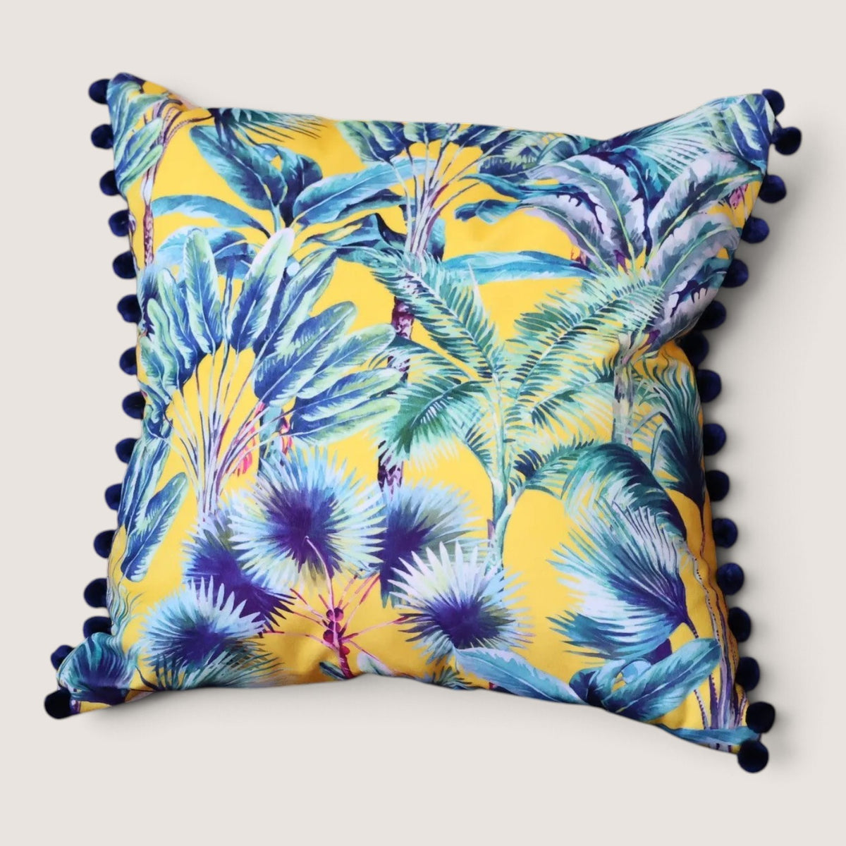 Palm Springs velvet cushion with navy pompoms, front view studio shot showing the yellow tropical palm and banana leaf print in teal, blue and purple, handmade in Dorset by Apricot Moon