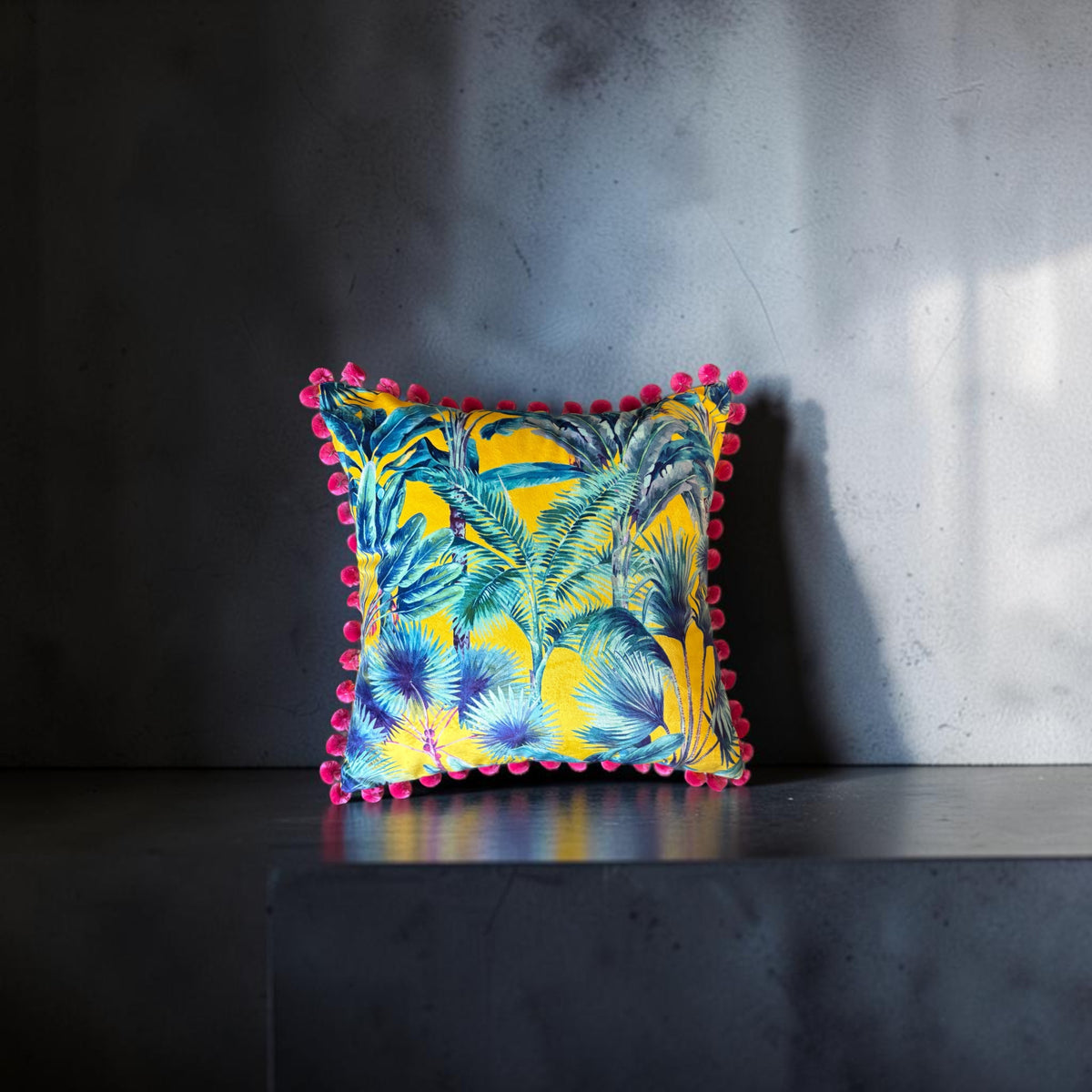 Palm Springs velvet cushion with strawberry pink pompoms, front view on a dark grey stand against a grey wall