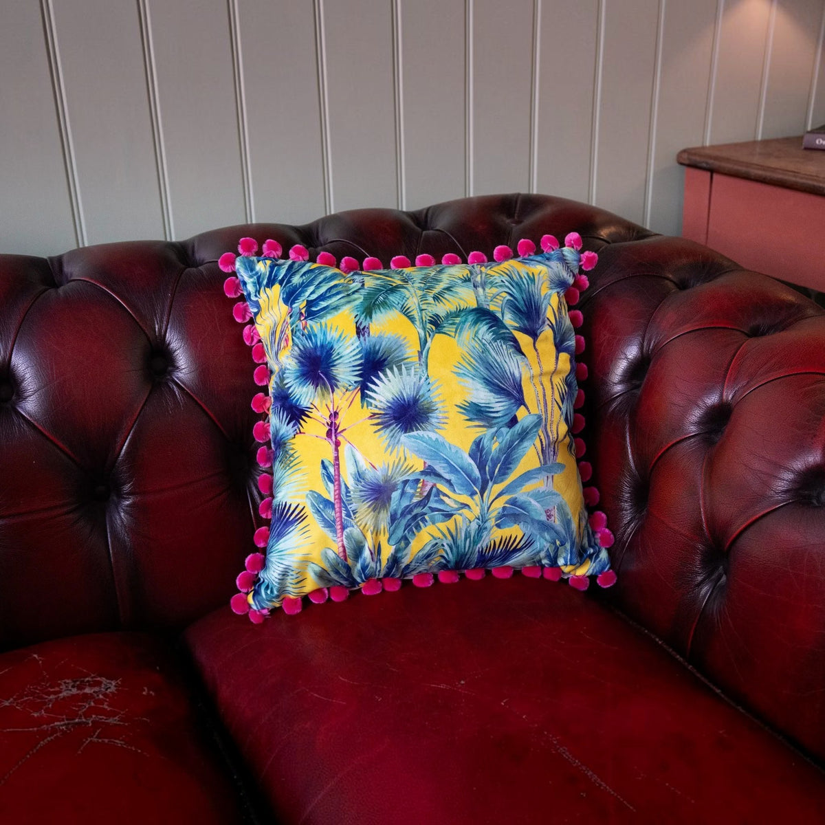 Palm Springs velvet cushion with strawberry pink pompoms on a deep red leather Chesterfield sofa