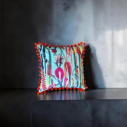 Paradise Blue velvet cushion with orange pompoms, front view on a dark grey stand against a grey wall