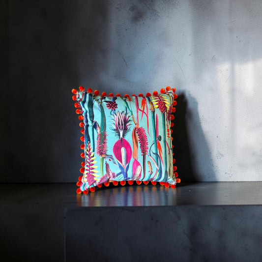 Paradise Blue velvet cushion with orange pompoms, front view on a dark grey stand against a grey wall