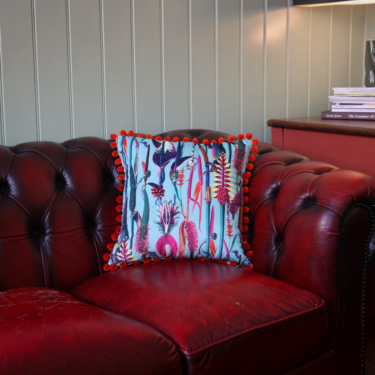 Paradise Blue velvet cushion with orange pompoms on a deep red leather Chesterfield sofa