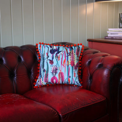 Paradise Blue velvet cushion with orange pompoms on a deep red leather Chesterfield sofa