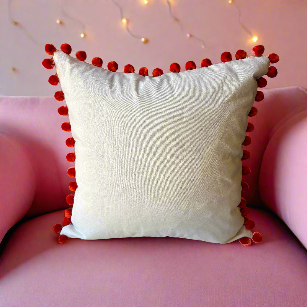 Rear view of the Paradise Blue cushion showing the plain cream back with orange pompoms, on a pink velvet sofa with fairy lights