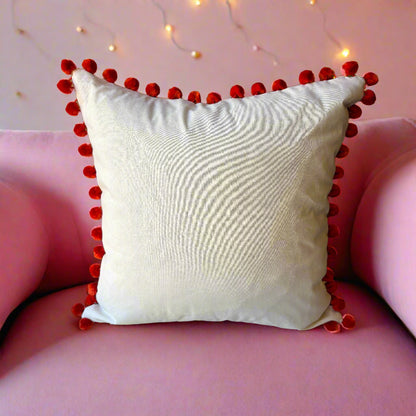 Rear view of the Paradise Blue cushion showing the plain cream back with orange pompoms, on a pink velvet sofa with fairy lights