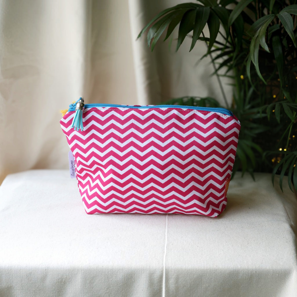 Raspberry Chevron everyday pouch in a pink and white zigzag print with a bright blue zip and pale blue tassel on a cream cloth beside a palm plant