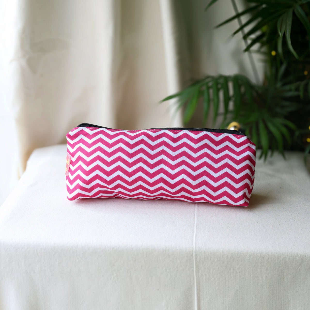 Raspberry Chevron pencil case in a pink and white zigzag print with a black zip on a cream cloth beside a palm plant