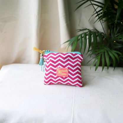 Raspberry Chevron coin purse with a turquoise zip and pale blue tassel on a cream cloth beside a palm plant