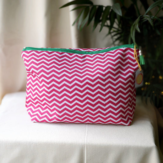 Raspberry Chevron wash bag in a pink and white zigzag print with a green zip on a cream cloth beside a palm plant