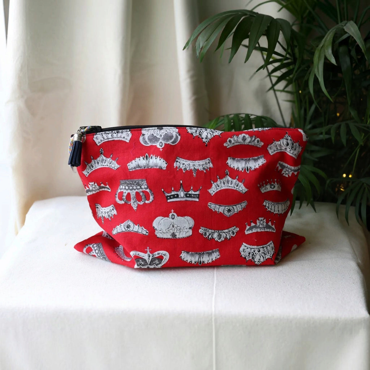 Red Crowns everyday pouch in red cotton printed with grey and white crowns and tiaras, front view on white cotton fabric by a green palm plant with twinkling lights, handmade in Dorset by Apricot Moon
