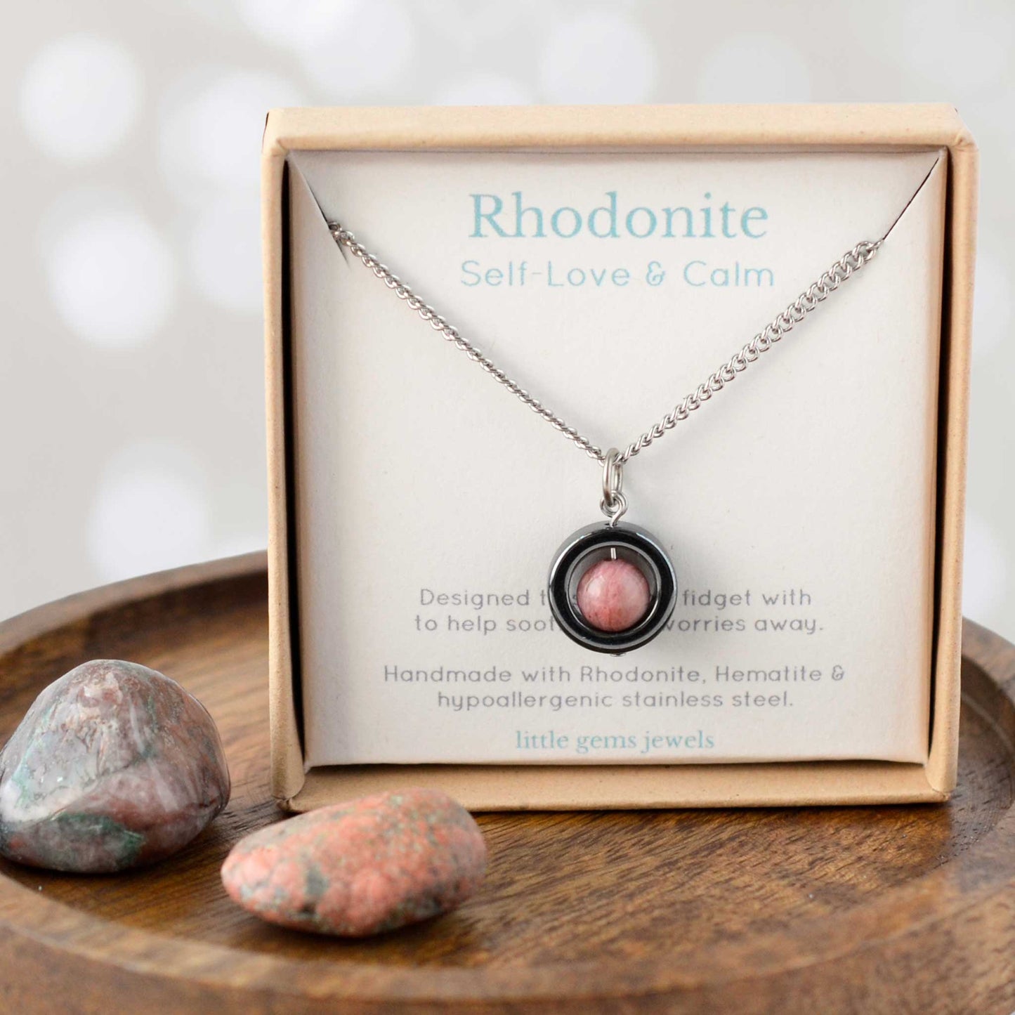Necklace with rhodonite pendant in a Little Gems Jewels box on a wooden surface with stones.