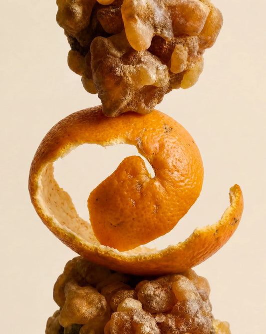 A close-up studio shot of a stacked fruit arrangement on a warm beige background: a curled orange peel ring around a small orange fruit, with clusters of amber-brown gummy or resin-like pieces above and below.