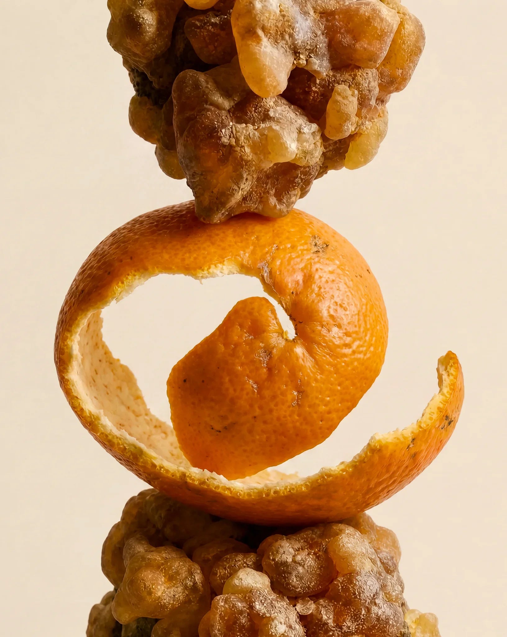 A close-up studio shot of a stacked fruit arrangement on a warm beige background: a curled orange peel ring around a small orange fruit, with clusters of amber-brown gummy or resin-like pieces above and below.