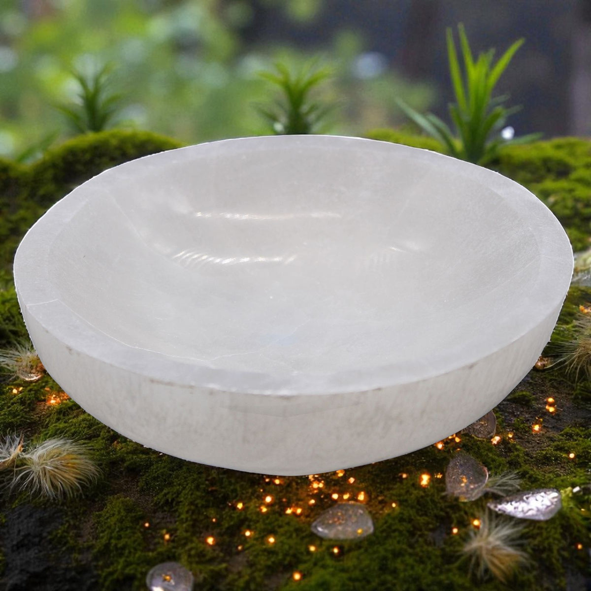 A white selenite bowl, placed on a bed of green moss with small fairy lights and decorative stones. The background is a soft-focus natural outdoor setting with green foliage.