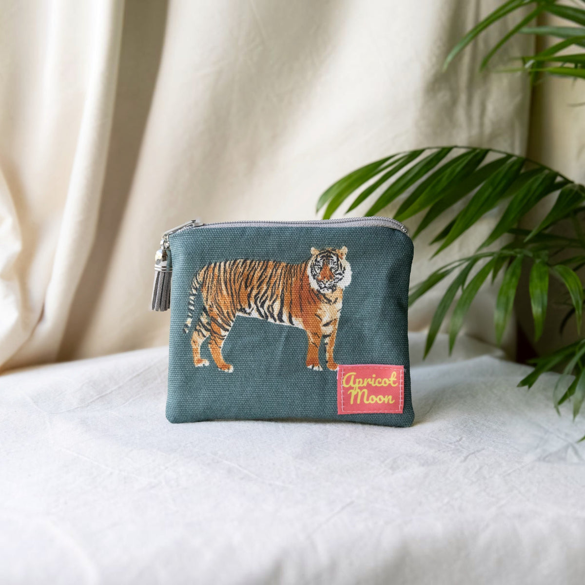 Tiger Tiger purse in a tiger print on deep teal with a grey zip and tassel, on a cream cloth beside a palm plant