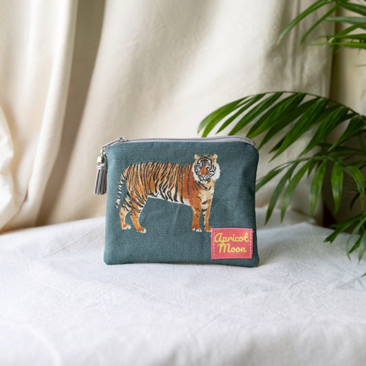 Tiger Tiger purse in a tiger print on deep teal with a grey zip and tassel, on a cream cloth beside a palm plant