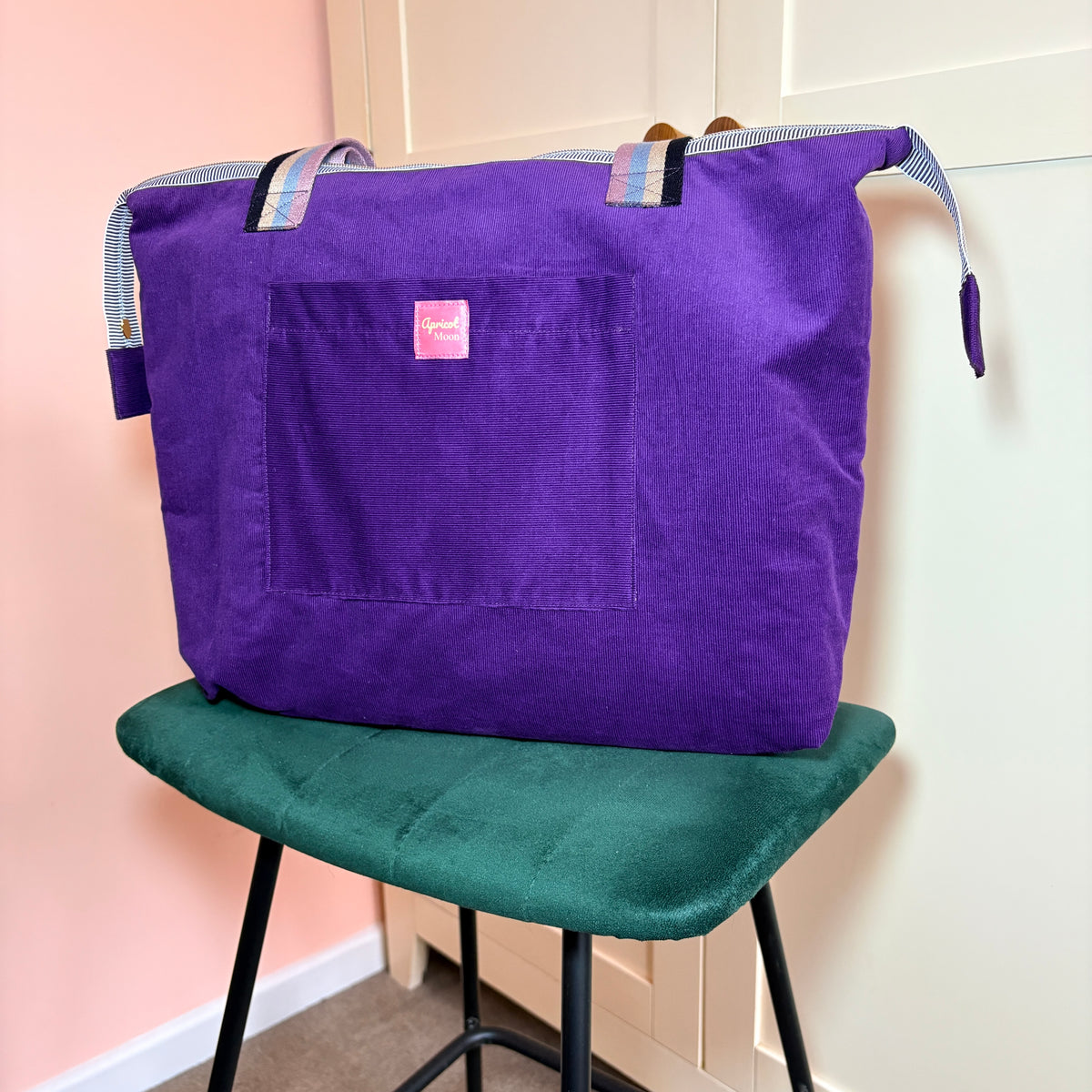 Twilight Bloom purple corduroy shoulder tote with striped handles and a front pocket, angled view on a green velvet stool