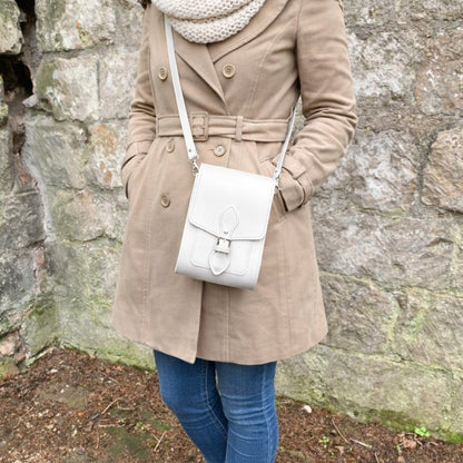 Person wearing a beige coat and blue jeans, holding a white handbag against a stone wall.