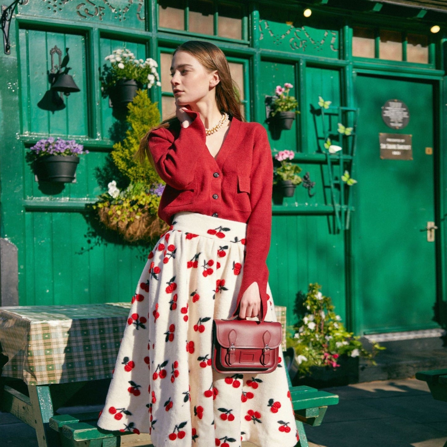 Woman in a red cardigan and cherry-patterned skirt standing in front of a green building with plants.